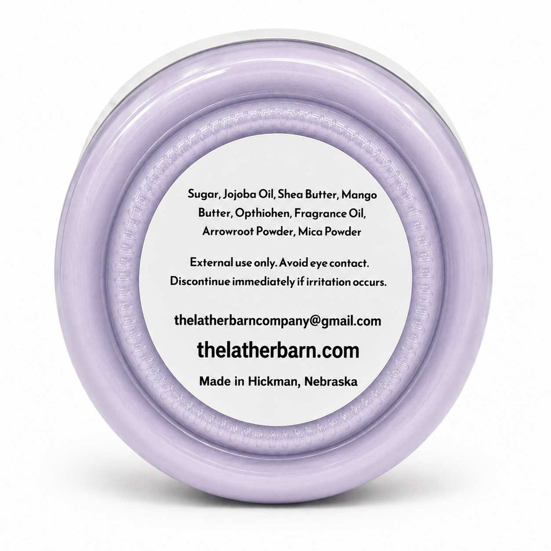 A round, clear plastic jar lid with a light lavender-purple rim, featuring a white circular label with black text listing ingredients and the website thelatherbarn.com.