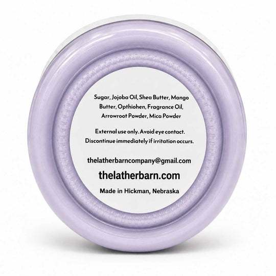 A round, clear plastic jar lid with a light lavender-purple rim, featuring a white circular label with black text listing ingredients and the website thelatherbarn.com.
