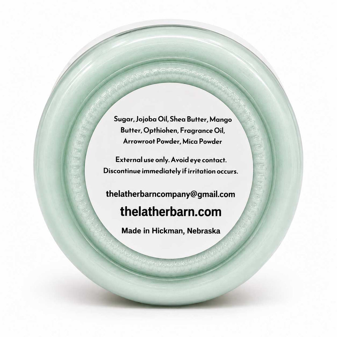 A round, clear plastic jar lid with a pale mint green tint, featuring a white circular label with black text listing ingredients and the brand 'The Lather Barn'.
