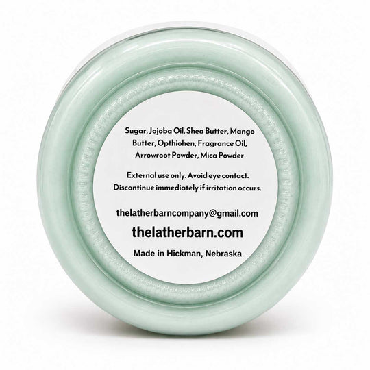 A round, clear plastic jar lid with a pale mint green tint, featuring a white circular label with black text listing ingredients and the brand 'The Lather Barn'.