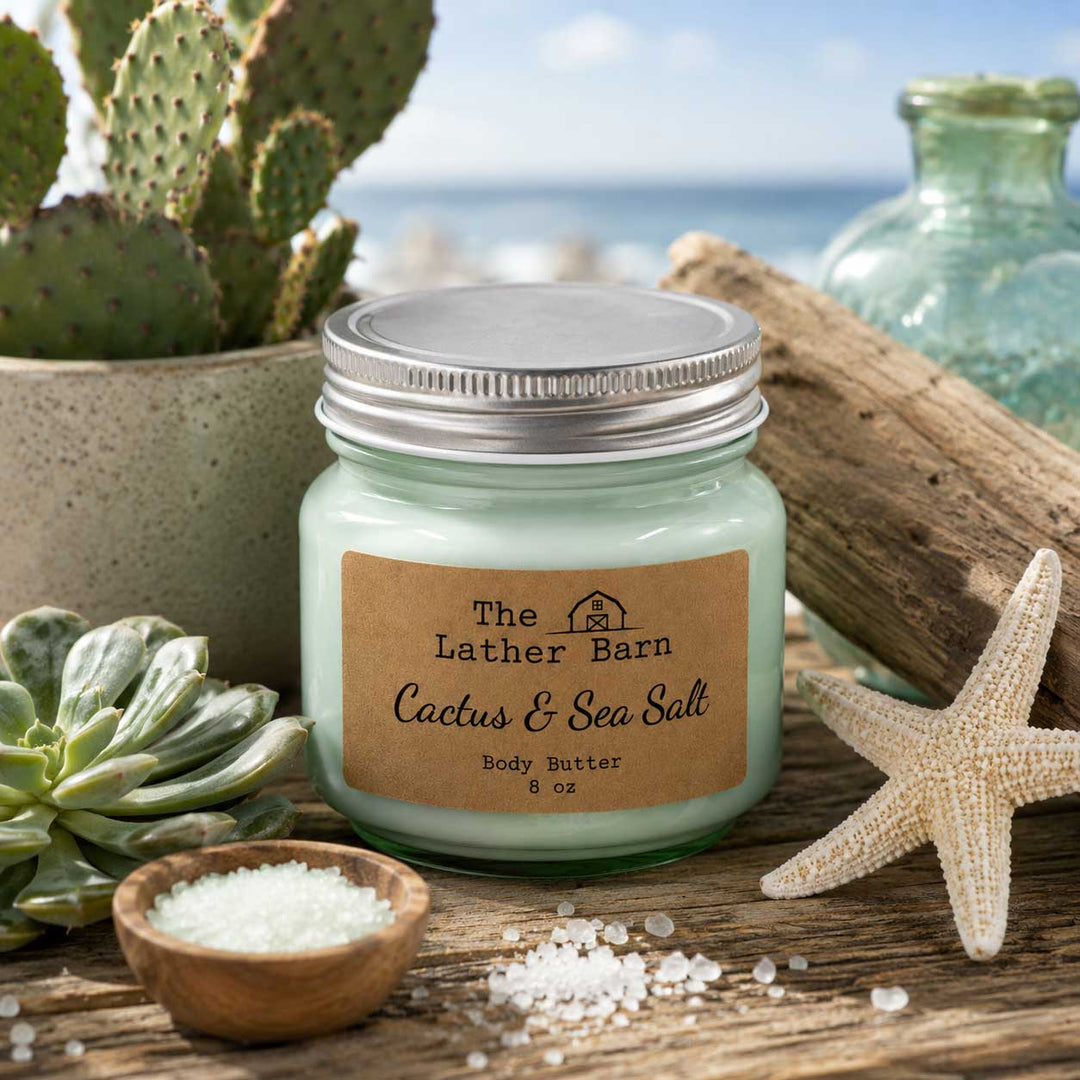 A jar candle labeled 'The Lather Barn Cactus & Sea Salt Body Butter' sits on a rustic wooden surface with beach-themed props including a starfish, small bowl of sea salt, cactus, succulent, driftwood, and ocean scenery in the background.