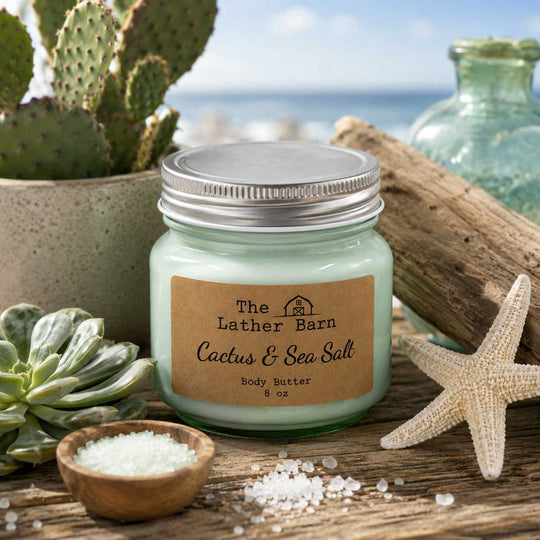 A jar candle labeled 'The Lather Barn Cactus & Sea Salt Body Butter' sits on a rustic wooden surface with beach-themed props including a starfish, small bowl of sea salt, cactus, succulent, driftwood, and ocean scenery in the background.