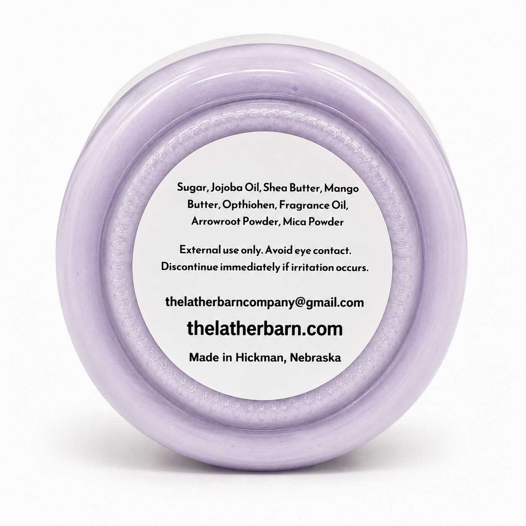 Top-down view of a round lavender-colored jar lid with a white label containing product ingredients and the brand name 'The Lather Barn'.