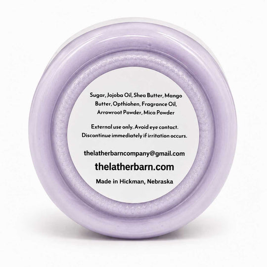Top-down view of a round lavender-colored jar lid with a white label containing product ingredients and the brand name 'The Lather Barn'.