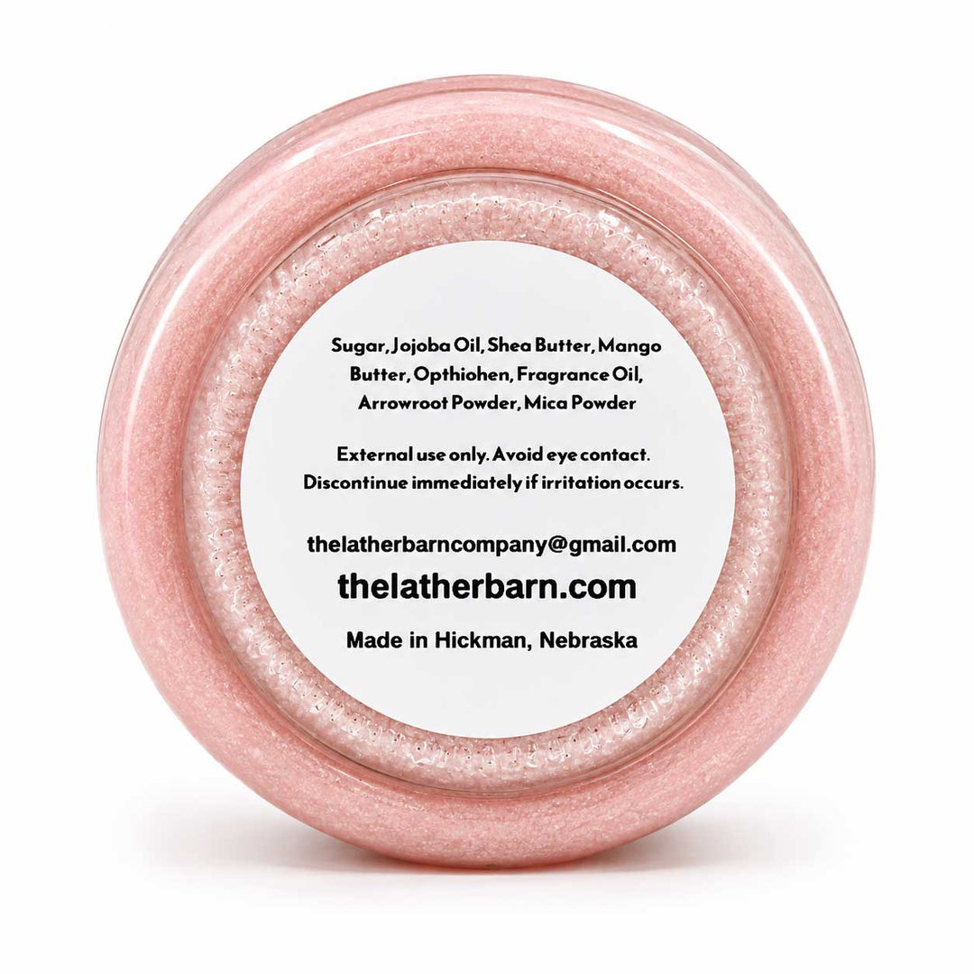 A round clear plastic jar filled with sparkling light pink cosmetic powder, featuring a white paper label with black text detailing the ingredients and brand 'The Lather Barn'.