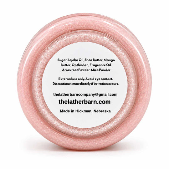 A round clear plastic jar filled with sparkling light pink cosmetic powder, featuring a white paper label with black text detailing the ingredients and brand 'The Lather Barn'.