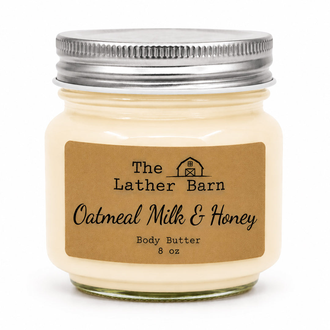 An 8 oz jar candle of 'The Lather Barn' featuring oatmeal milk and honey body butter. The candle is in a clear glass jar filled with creamy off-white wax, topped with a silver metal lid, and has a brown kraft paper label.