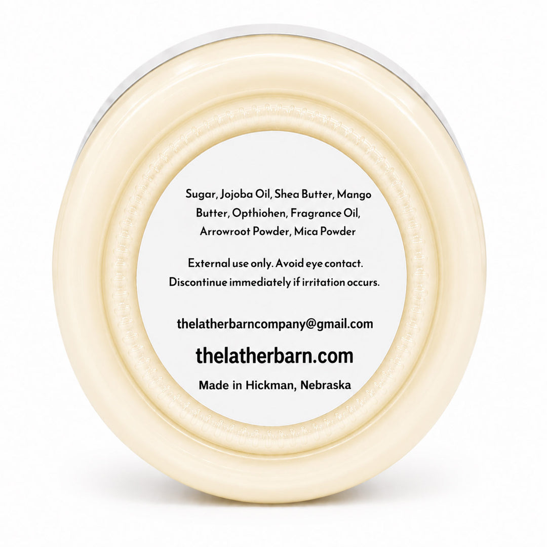 Top-down view of a round cream-colored jar lid with a white center label containing product ingredients and the brand name 'thelatherbarn.com'.