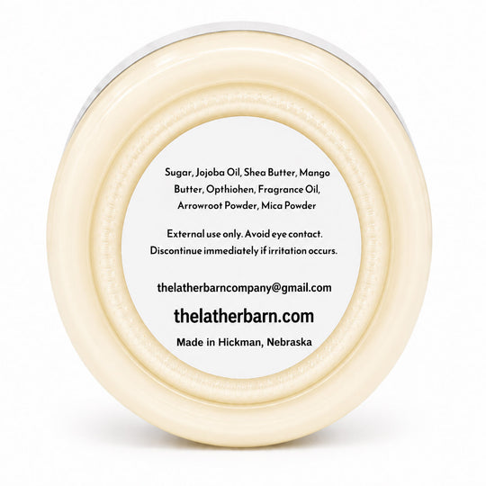 Top-down view of a round cream-colored jar lid with a white center label containing product ingredients and the brand name 'thelatherbarn.com'.
