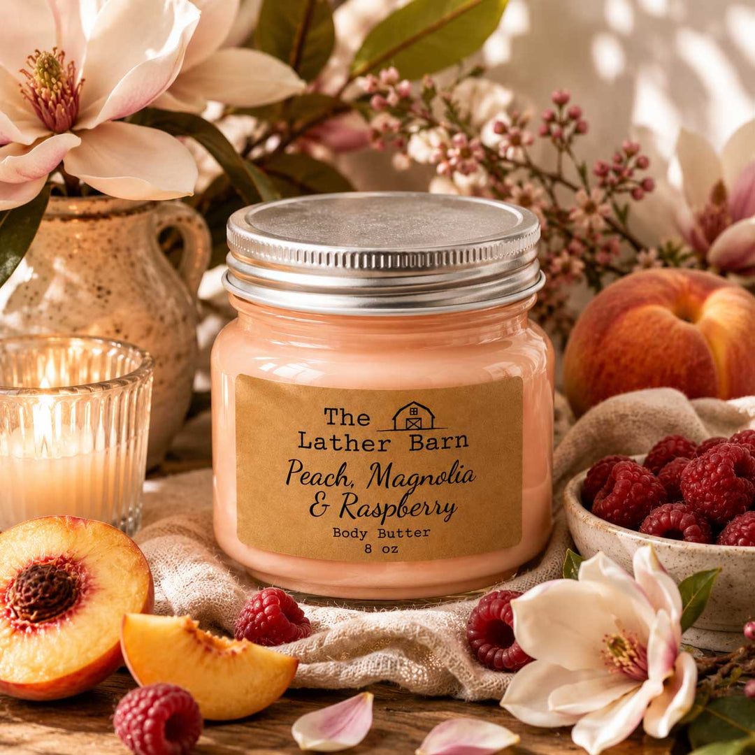 A jar of peach, magnolia, and raspberry body butter by The Lather Barn sits on a rustic wooden surface surrounded by peaches, raspberries, white flowers, and a lit candle. The product is in a clear glass jar with a silver lid and a tan paper label.