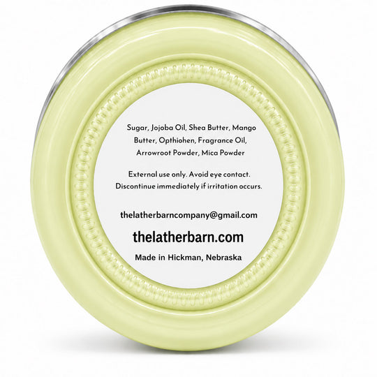 Top-down view of a round pale yellow cosmetic jar with a clear lid and a white label showing product ingredients and the brand thelatherbarn.com.