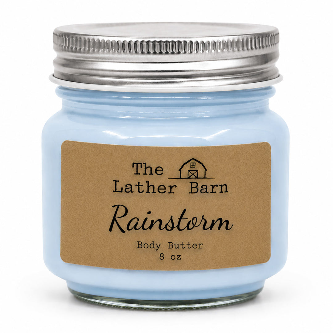 A light blue glass jar candle with a silver metal lid and a brown kraft paper label reading 'The Lather Barn Rainstorm Body Butter 8 oz' on a white background.