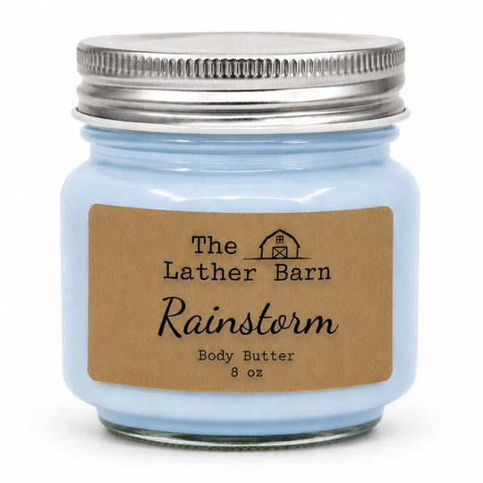 A light blue glass jar candle with a silver metal lid and a brown kraft paper label reading 'The Lather Barn Rainstorm Body Butter 8 oz' on a white background.