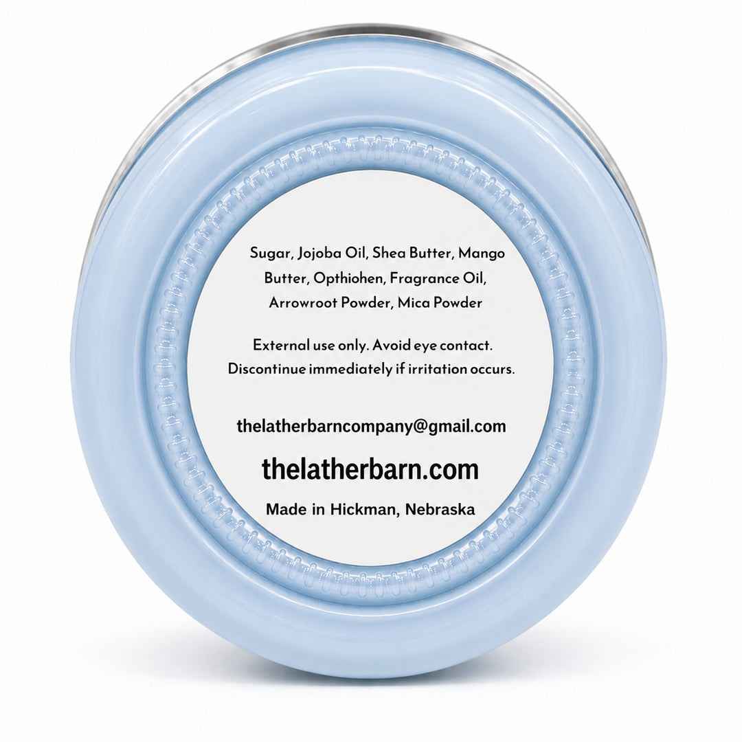 A light blue glass jar candle with a silver metal lid and a brown kraft paper label reading 'The Lather Barn Rainstorm Body Butter 8 oz' on a white background.