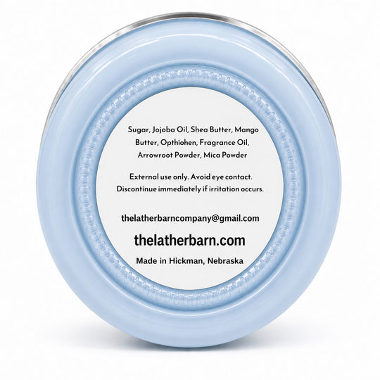 A light blue glass jar candle with a silver metal lid and a brown kraft paper label reading 'The Lather Barn Rainstorm Body Butter 8 oz' on a white background.