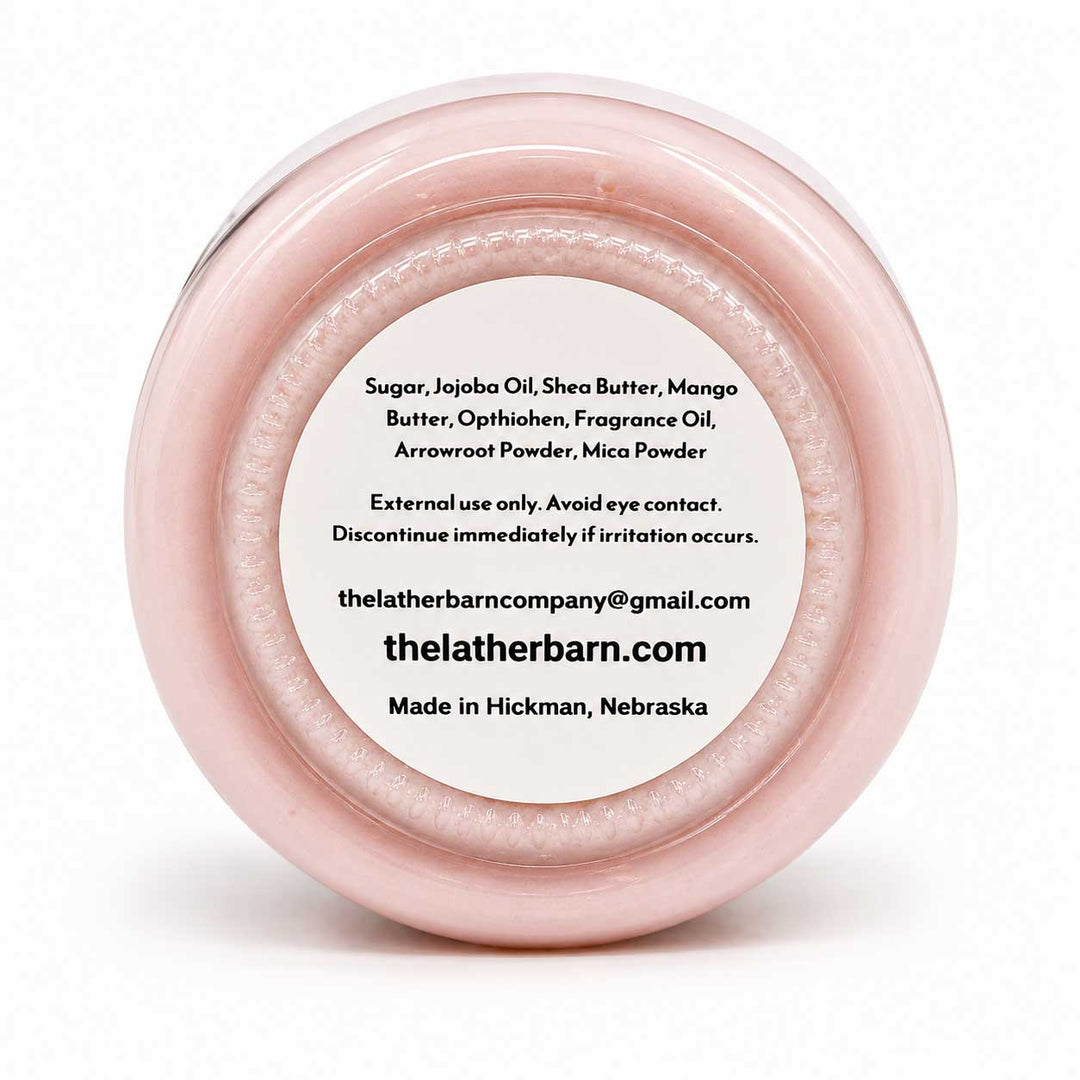 A round, open jar of Thelather Barn Company sugar scrub with a light pink translucent lid and a white label showing product ingredients and contact information, set against a plain white background.