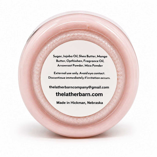 A round, open jar of Thelather Barn Company sugar scrub with a light pink translucent lid and a white label showing product ingredients and contact information, set against a plain white background.