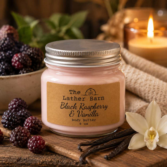 A jar candle labeled 'The Lather Barn Black Raspberry & Vanilla Body Butter' sits on a rustic wooden surface with blackberries, vanilla beans, a white orchid flower, and a lit candle in the background.