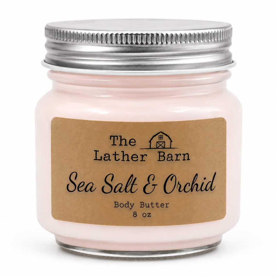 A jar of Sea Salt & Orchid body butter by The Lather Barn, featuring a light pink cream inside a clear glass jar with a silver metal lid and a brown kraft paper label.