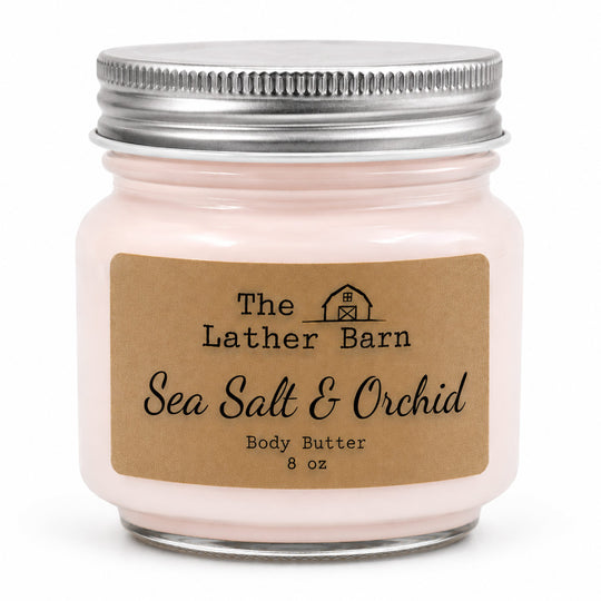 A jar of Sea Salt & Orchid body butter by The Lather Barn, featuring a light pink cream inside a clear glass jar with a silver metal lid and a brown kraft paper label.