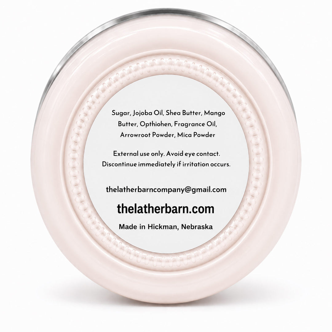 Top-down view of a round, pale pink cosmetic jar lid with a silver metal rim and a white label containing product ingredients and the brand name 'The Lather Barn'.