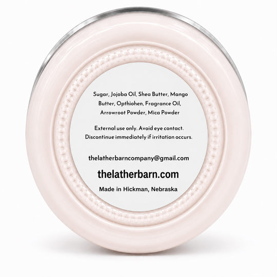 Top-down view of a round, pale pink cosmetic jar lid with a silver metal rim and a white label containing product ingredients and the brand name 'The Lather Barn'.