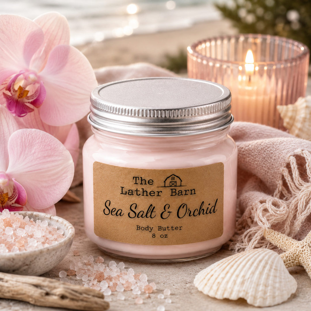 A jar of 'The Lather Barn' Sea Salt & Orchid body butter is styled on a beach-themed surface with pink orchids, a lit candle, seashells, and sea salt crystals.