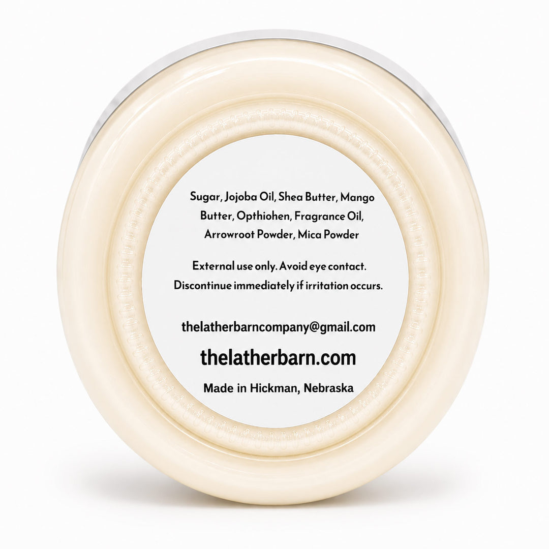 A round, off-white cosmetic jar viewed from above, showing a white label with black text listing ingredients and the brand name 'thelatherbarn.com'.