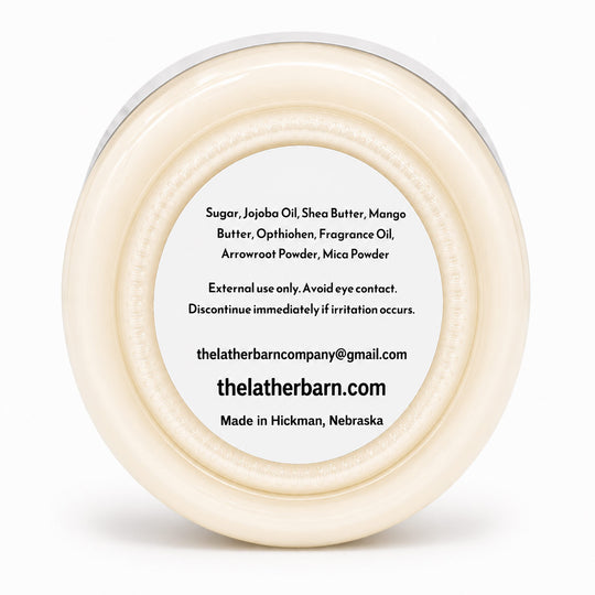 A round, off-white cosmetic jar viewed from above, showing a white label with black text listing ingredients and the brand name 'thelatherbarn.com'.