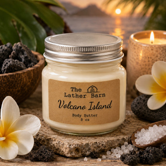A jar of body butter from The Lather Barn, labeled 'Volcano Island', styled on a rustic stone coaster with lava rocks, white flowers, and a warm candle in the background.