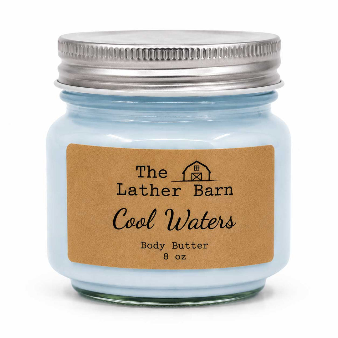 A light blue glass jar candle with a silver metal lid and a brown kraft paper label reading 'The Lather Barn Cool Waters Body Butter 8 oz' on a white background.