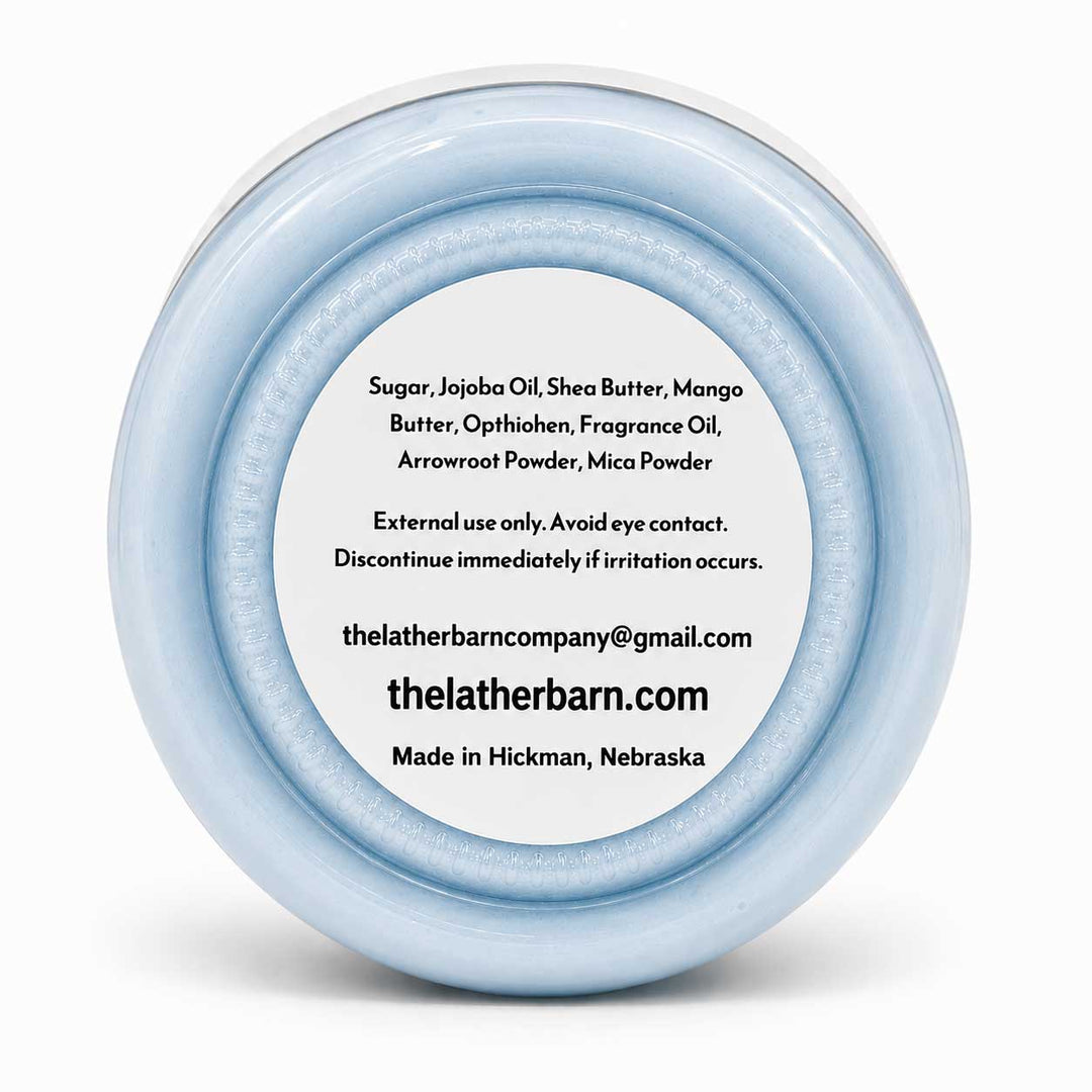 A round, light blue cosmetic jar lid with a white center label displaying product ingredients and the brand name 'The Lather Barn'.