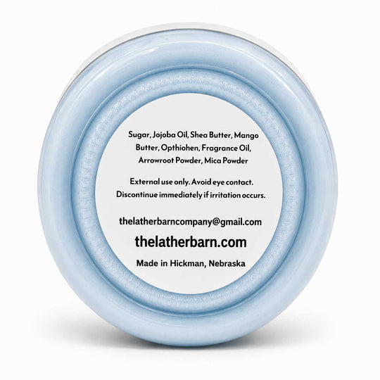 A round, light blue cosmetic jar lid with a white center label displaying product ingredients and the brand name 'The Lather Barn'.
