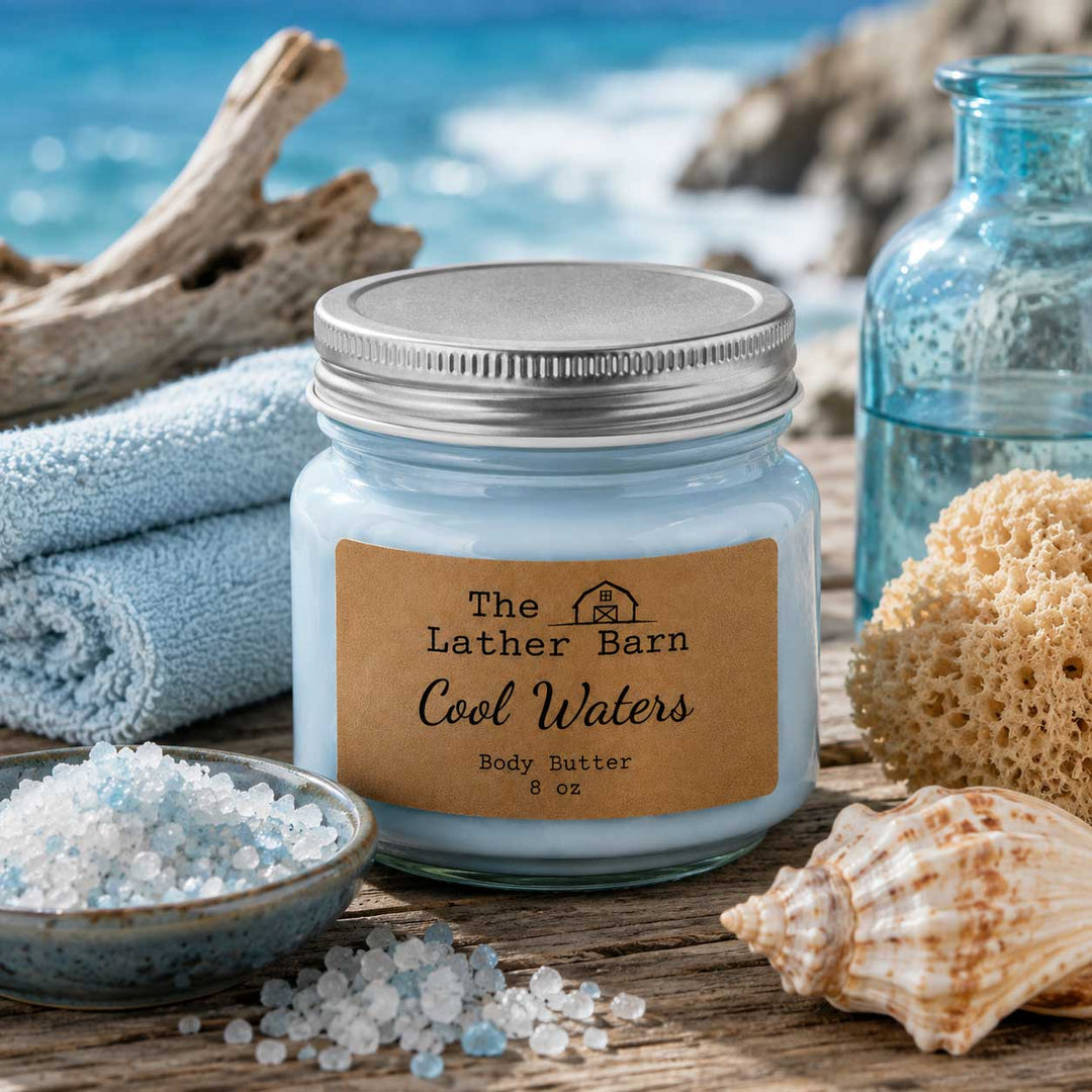 A jar candle labeled 'The Lather Barn Cool Waters Body Butter 8 oz' sits on a wooden surface with beach-themed props including a towel, seashells, a bowl of bath salts, driftwood, and a blue glass bottle against a blurred ocean background.