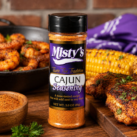 Bottle of Misty's Cajun seasoning with grilled chicken, corn, and herbs on a wooden surface.