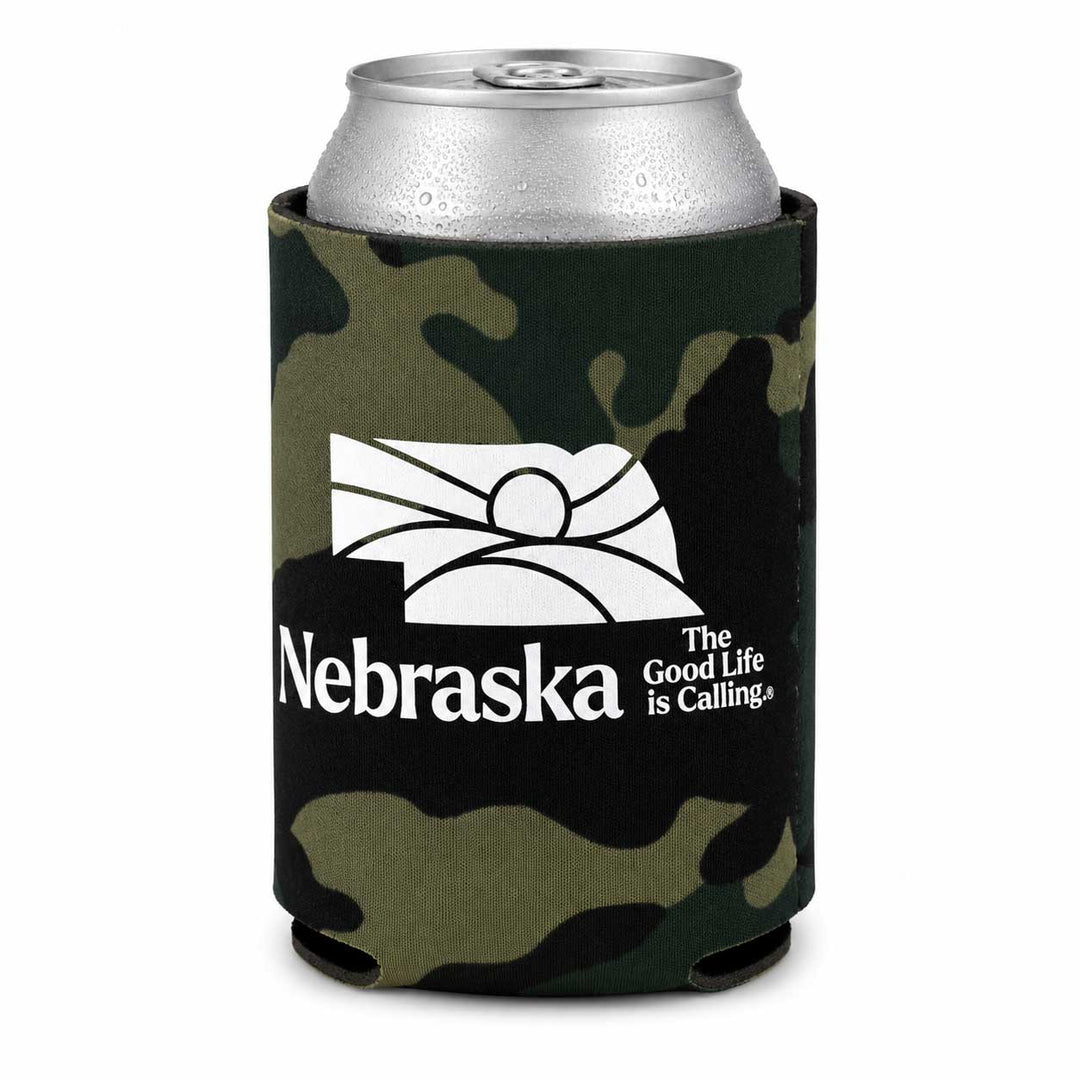 A can cooler sleeve with a green and black camouflage pattern, featuring a white printed graphic of the Nebraska state silhouette with a rising sun and the text 'Nebraska The Good Life is Calling'. A silver aluminum can is inserted into the sleeve, shown against a plain white background.
