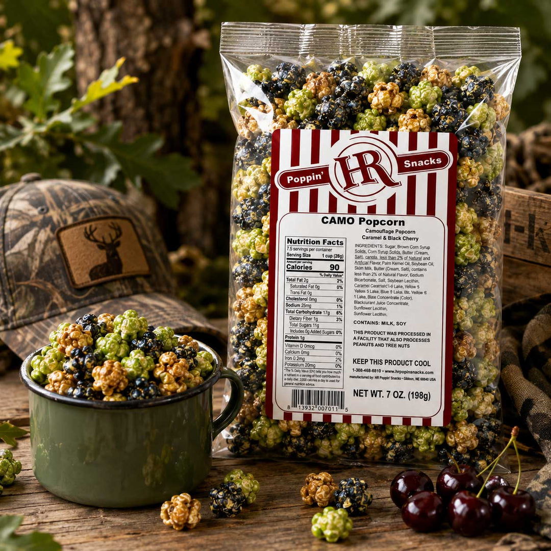 A bag of Poppin' HR Camo Popcorn stands on a rustic wooden surface with a green enamel bowl filled with popcorn in front of it. The popcorn kernels are mixed green, black, and tan in a camouflage pattern, matching the woodland-themed props including a camouflage hat and cherries.