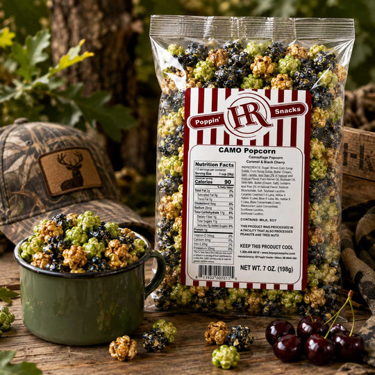 A bag of Poppin' HR Camo Popcorn stands on a rustic wooden surface with a green enamel bowl filled with popcorn in front of it. The popcorn kernels are mixed green, black, and tan in a camouflage pattern, matching the woodland-themed props including a camouflage hat and cherries.
