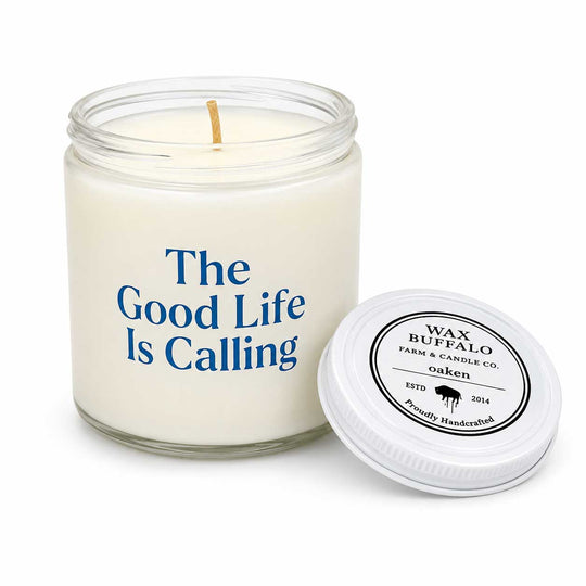 A white soy wax candle in a clear glass jar with blue text reading 'The Good Life Is Calling,' shown next to its white screw-top lid on a white background. The lid label reads 'Wax Buffalo Farm & Candle Co.