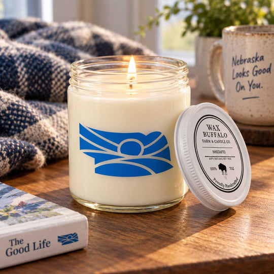 A lit candle in a clear glass jar sits on a wooden table with its white lid placed beside it. The jar has a blue abstract logo, and the lid reads 'WAX BUFFALO YARN & CASTLE CO.