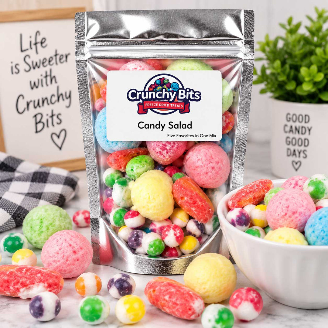 A stand-up pouch of Crunchy Bits Candy Salad filled with colorful hard candies, shown on a marble surface with a bowl and scattered candies, plus decorative signs and a plant in the background.