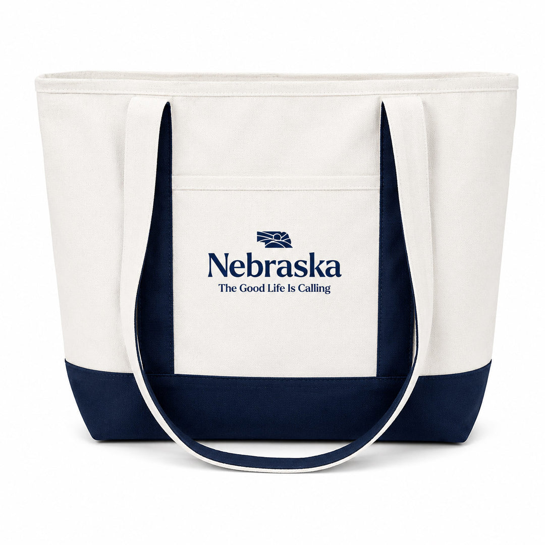 A white canvas tote bag with dark navy blue trim and handles, featuring a front pocket printed with the Nebraska logo and the text 'The Good Life Is Calling'.