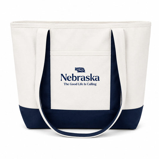 A white canvas tote bag with dark navy blue trim and handles, featuring a front pocket printed with the Nebraska logo and the text 'The Good Life Is Calling'.