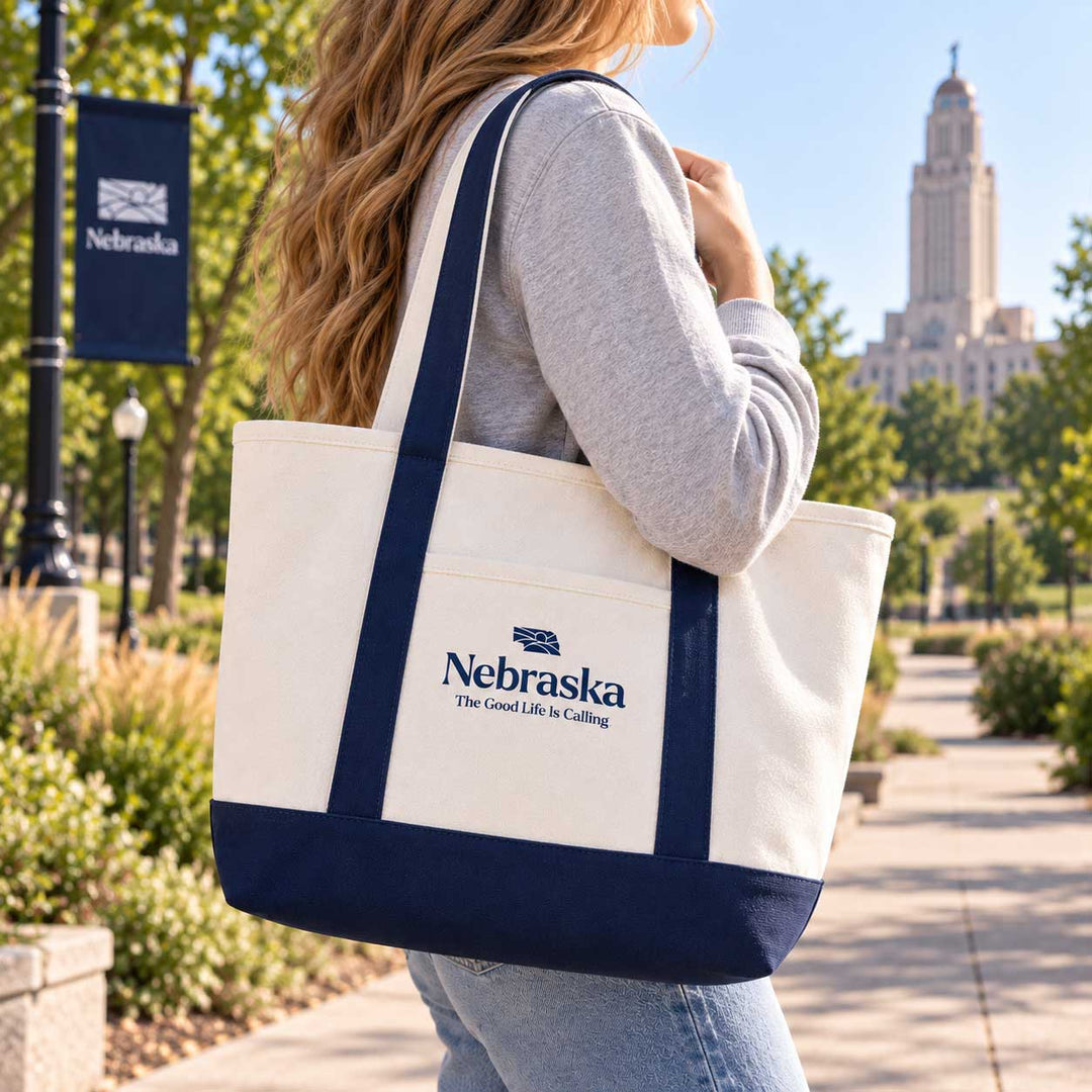 A woman carrying a large cream canvas tote bag with navy blue handles and bottom trim. The tote bag features a front pocket printed with the Nebraska state logo and the text 'Nebraska The Great State of Mind'.