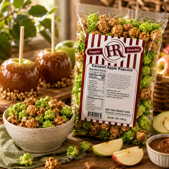 A bag of Poppin' Snacks Caramel Apple Popcorn stands on a rustic wooden table with a bowl of the popcorn in front and caramel-apple props in the background. The packaging is clear plastic with a large red-and-white striped label, and visible popcorn pieces and apple slices are scattered around.