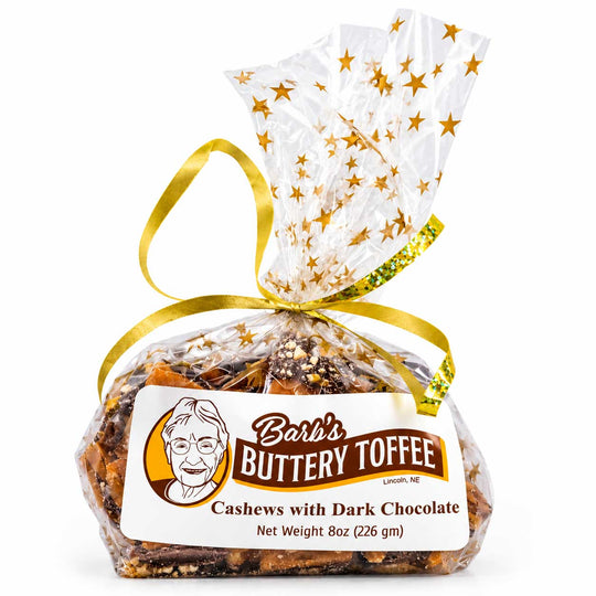 A 8 oz bag of Barb's Buttery Toffee Cashews with Dark Chocolate, sealed in clear cellophane with a gold ribbon and a white top section decorated with gold stars, on a white background.