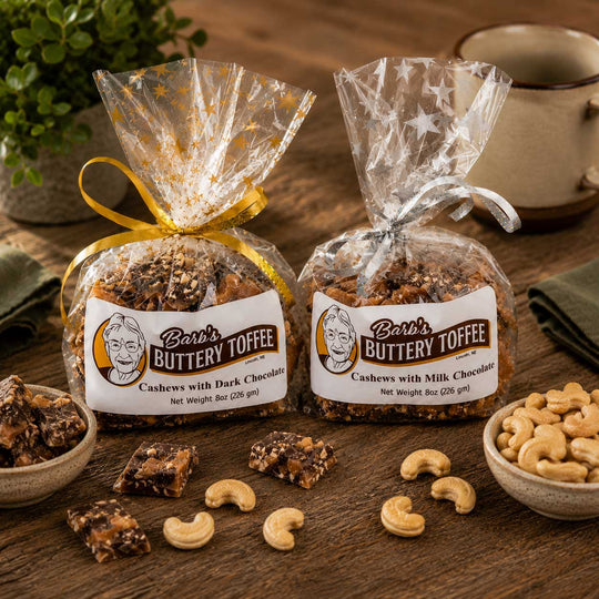 Two bags of Barb's Buttery Toffee, one with dark chocolate and one with milk chocolate, wrapped in clear cellophane and tied with ribbons, displayed on a wooden table with cashews and toffee pieces.