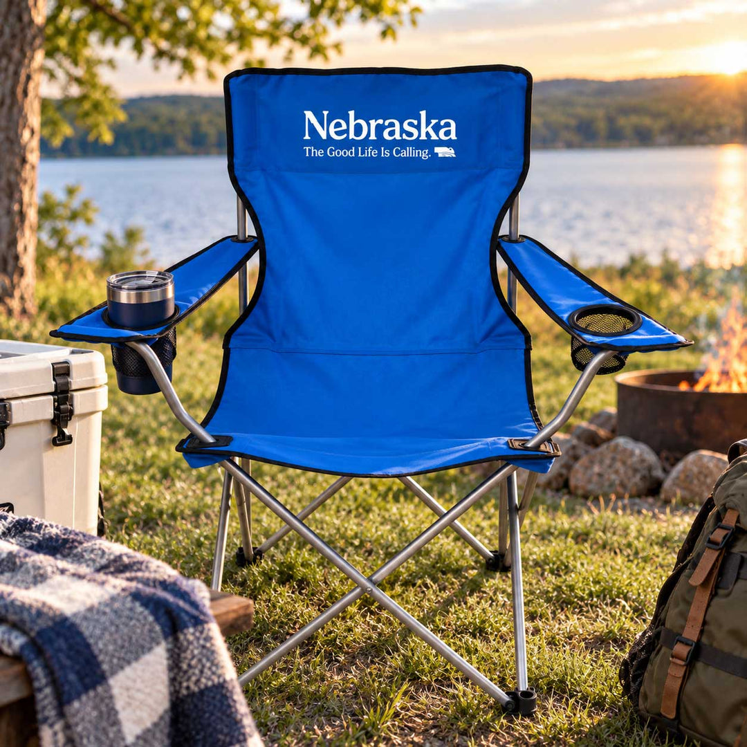 A blue folding camp chair with 'Nebraska The Good Life Is Calling.' printed on the back, set on grass by a lake at sunset with a cooler, blanket, backpack, and fire pit visible.