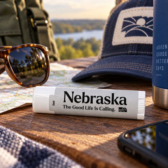 A white lip balm tube labeled 'Nebraska The Good Life Is Calling.' lies on a wooden table outdoors, surrounded by sunglasses, a blue cap, a map, a blue bottle, and a phone.