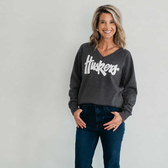 A woman wearing a dark gray v-neck sweatshirt with 'Huckers' printed in white script across the chest, paired with dark blue jeans, standing against a light gray wall.